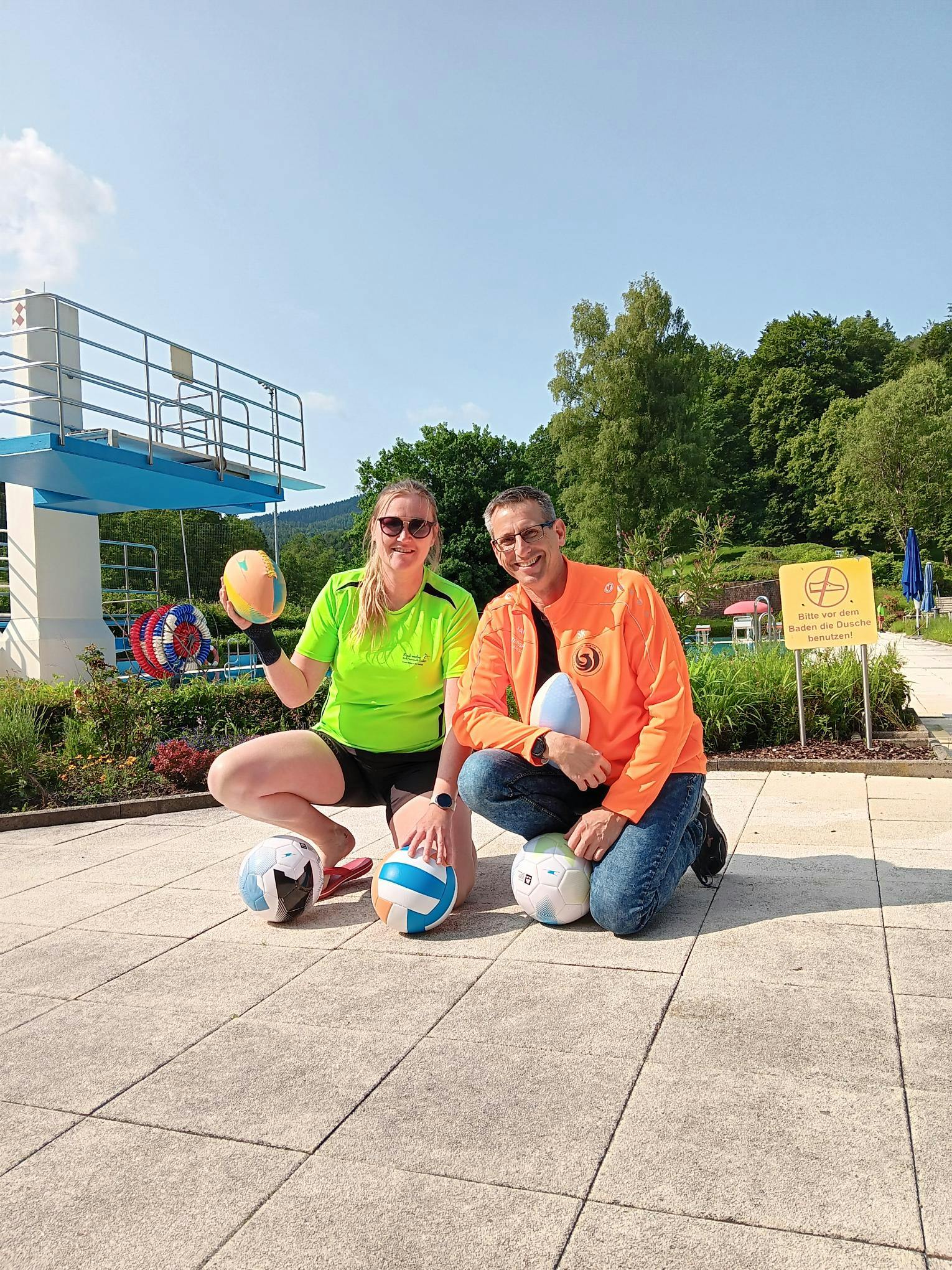 Frau Christiane Kühn (Waldfreibad-Leitung) und Sascha Reddig (Phönix Albtal e.V.) bei der Übergabe der Sportgeräte: gut gelaunt im Freibad, umgeben von Bällen und Sommerlaune.